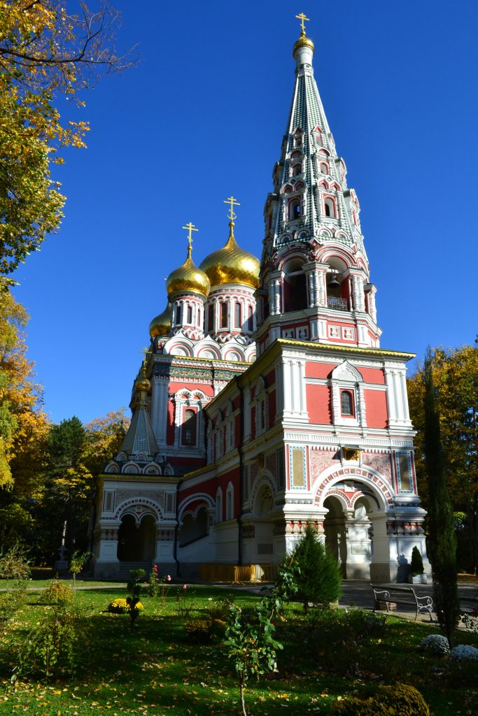 Large Russian Orthodox church with soft pink highlights and golden onion domes situated in a lush garden with grass and leafy trees with their fall colors